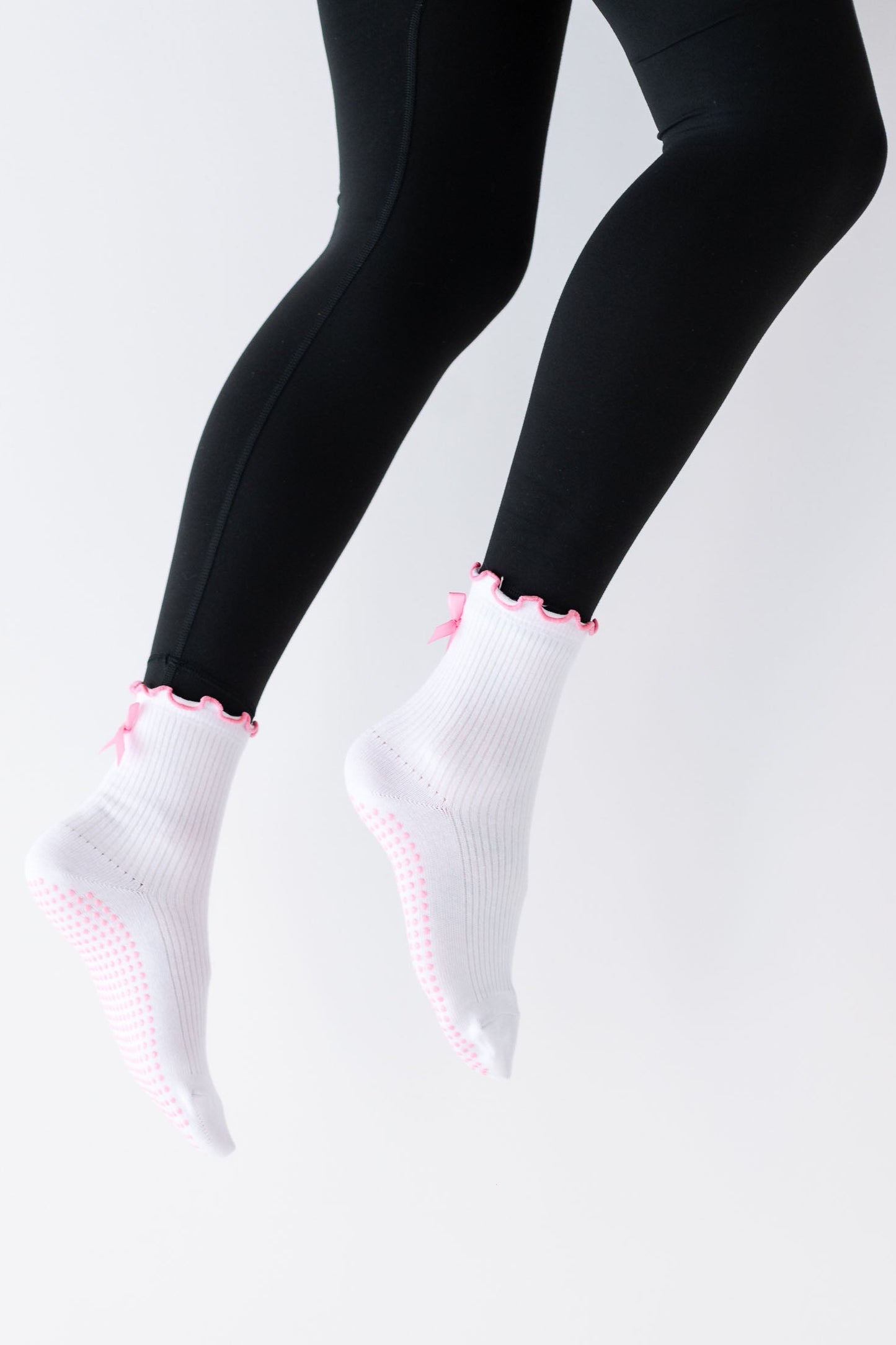 A pair of white ribbed crew socks with a pink bow detail on the heel, worn on legs raised in the air against a white background.