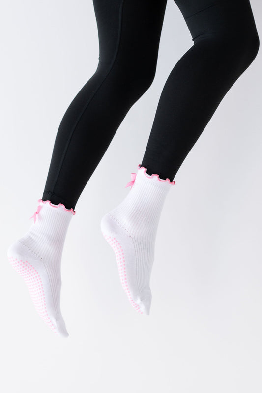 A pair of white ribbed crew socks with a pink bow detail on the heel, worn on legs raised in the air against a white background.