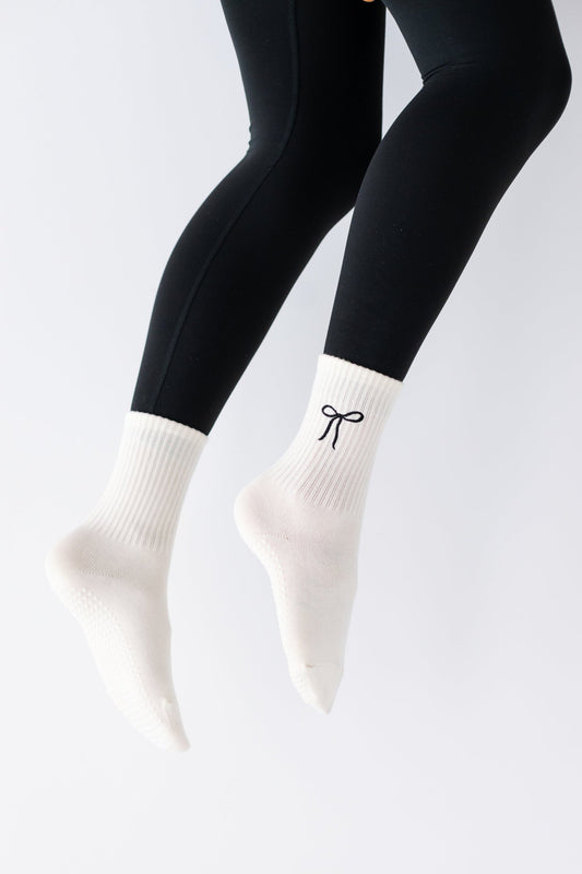 A pair of beige 'Sealed with a Bow' crew socks with a bow design on the ankle area, worn with black leggings.